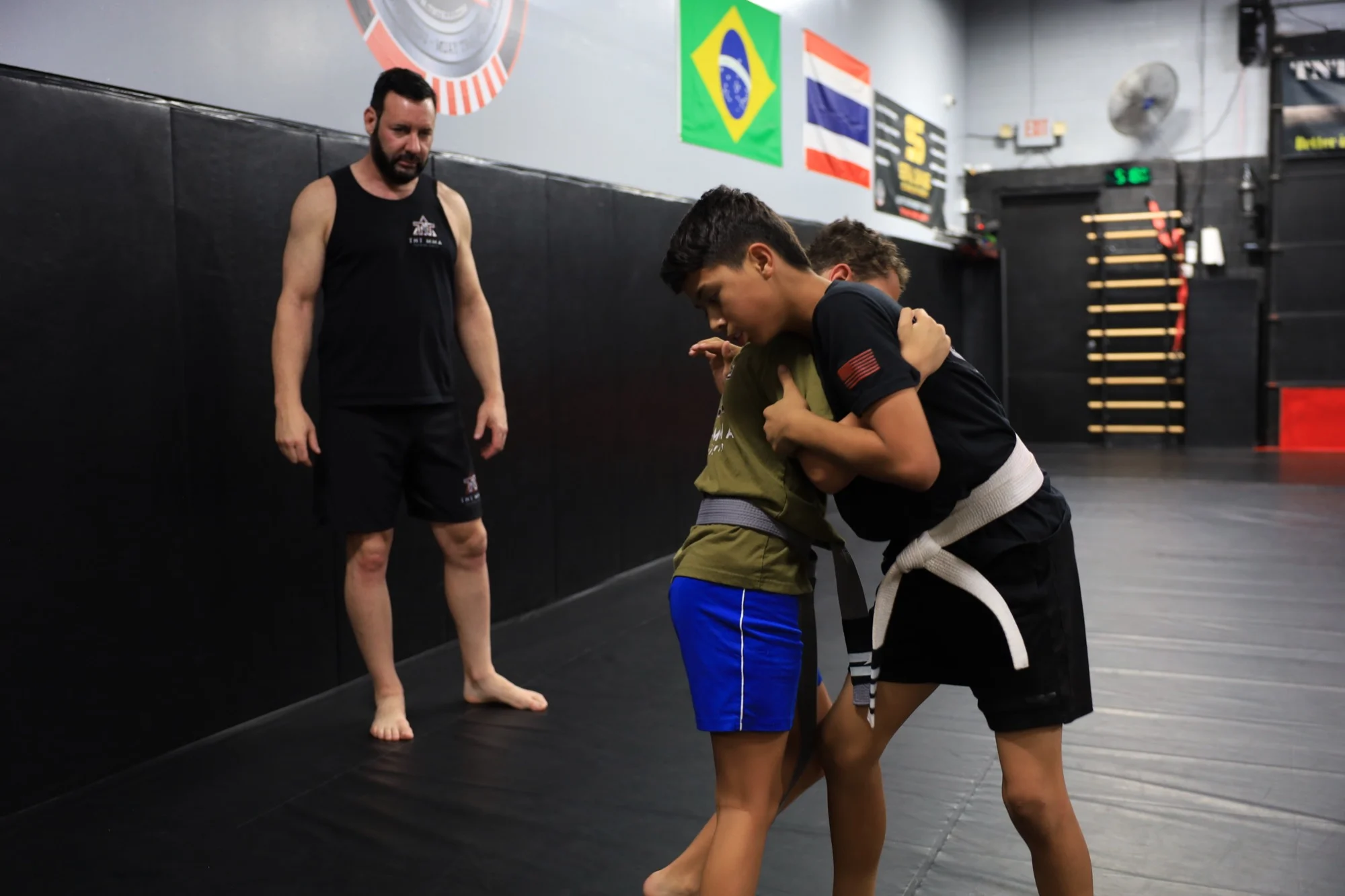 A TNT MMA coach supervising two kids practicing clinch work in a youth MMA class in Phoenix, Arizona